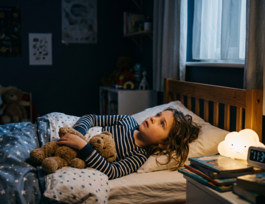 Child lying in bed hugging a teddy bear with a nightlight and clock nearby